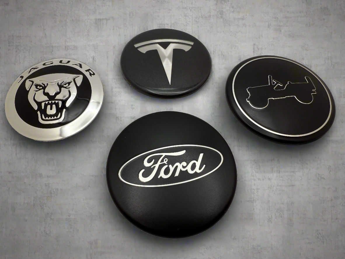 Four car brand logo emblems on a gray surface. Brands include Ford, Jeep, Tesla & Jaguar. 