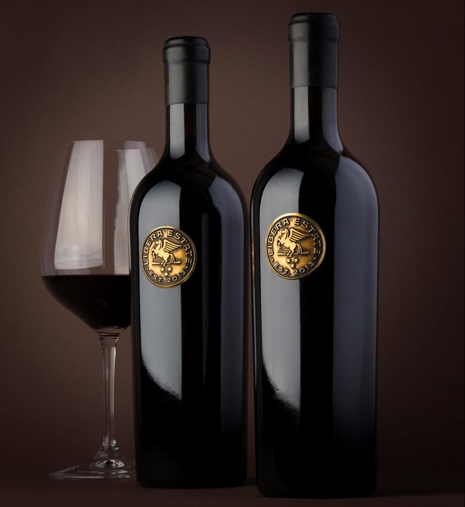 2 black wine bottles next to a glass of half full wine. Wine bottles have circular antique bronze metal labels attached